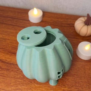 May include: A light green pumpkin-shaped container with a lid, next to two lit candles and a small pumpkin. The container has a round opening and a decorative design. The candles and pumpkin add to the fall theme.