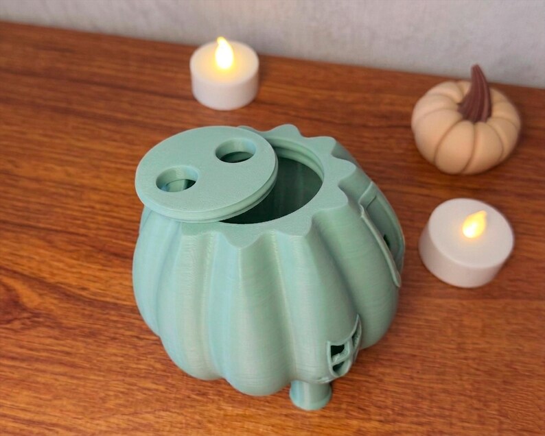 May include: A light green pumpkin-shaped container with a lid, next to two lit candles and a small pumpkin. The container has a round opening and a decorative design. The candles and pumpkin add to the fall theme.