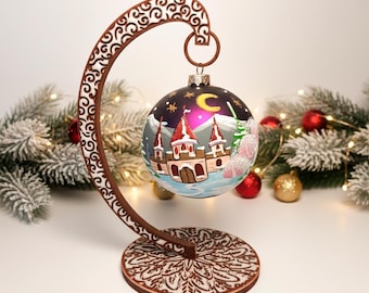May include: A decorative Christmas ornament featuring a winter castle scene with red-roofed towers, a crescent moon, and stars. The ornament hangs from a brown, ornate stand with a circular base. Festive red and gold ornaments and greenery are in the background.