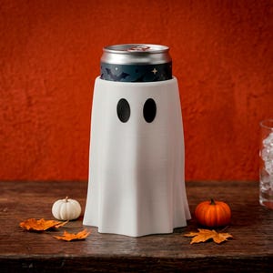 May include: A white ghost-shaped beverage can cooler featuring two black oval eyes. The cooler holds a silver aluminum can with a dark blue label decorated with black bat silhouettes and small gold stars, creating a festive seasonal appearance.