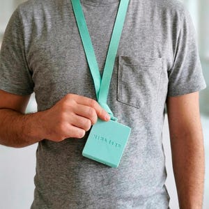 Op de afbeelding: Een lichtblauwe lanyard met een bijpassend vierkant badge. Op de badge staat de tekst "MiRAdsgn" in reliëf. De lanyard hangt om de nek van een persoon die een grijs t-shirt draagt.
