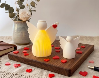 May include: Two angel-shaped decorative lights, one illuminated with a warm glow, sit on a wooden tray. The angels have white wings and a red heart detail. Red and pink heart-shaped decorations are scattered around the angels.