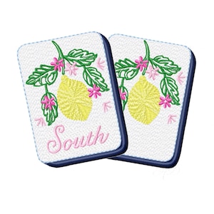 May include: Two white coasters with a navy blue border. Each coaster features a yellow lemon, green leaves, and pink flowers. The word "South" is written in pink script on one coaster.