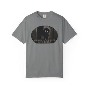 May include: Gray t-shirt with a black and white graphic of a creature peeking from behind a tree in a forest. The text "WANNA PLAY HIDE AND SEEK?" is printed below the graphic. The shirt has short sleeves and a crew neck.