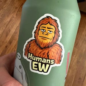 May include: A green water bottle with a sticker featuring a cartoon Sasquatch with the text "Humans EW". The sticker has a white border and the Sasquatch is brown and orange.