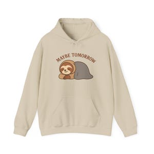 May include: Beige hoodie with a kangaroo pocket and a drawstring hood. The front features a cartoon sloth illustration with the text "MAYBE TOMORROW" in brown. The sloth is brown and gray, and the hoodie is a neutral color.
