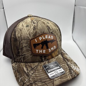 May include: A brown and tan camouflage trucker hat with a brown patch that says "I Plead The 2nd" with a silhouette of a rifle. The hat is adjustable and has a black mesh back.