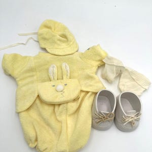 May include: A yellow baby outfit set, including a romper with bunny ears, a matching hat, a pair of booties, and two gray shoes. The romper has a bunny face design on the front. The set is made of soft material.