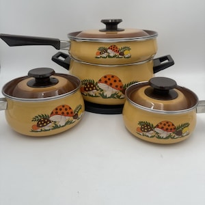 Peut inclure: Un ensemble de trois ustensiles de cuisine en émail jaune avec des couvercles marron et un motif de champignons. L'ensemble comprend une grande casserole avec un long manche et deux petites marmites avec des poignées latérales.