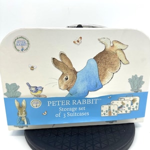 May include: A cream-colored Peter Rabbit storage set of 3 suitcases. The suitcases feature illustrations of Peter Rabbit, butterflies, bees, and a blue bird. The set has a handle and a blue banner with the text "PETER RABBIT Storage set of 3 Suitcases."