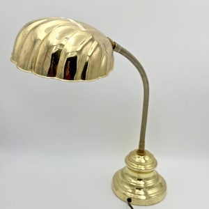 Peut inclure: Une lampe de bureau en laiton poli dotée d'un abat-jour festonné en forme de coquillage. La lampe possède un bras flexible de type col de cygne relié à une base ronde à gradins. Un cordon électrique noir sort de la base au fini doré métallique.