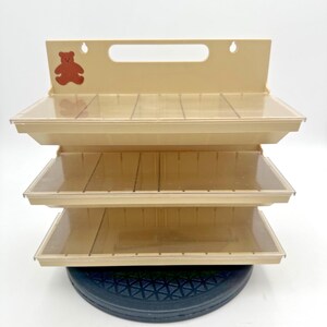May include: A beige plastic wall-mounted storage organizer featuring three horizontal shelves with clear, hinged lids. The top panel includes a cutout handle, two mounting holes, and a small brown bear graphic on the left side.