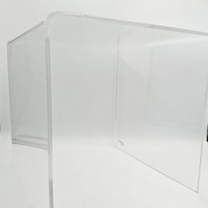May include: A clear acrylic side table with a minimalist design. The table has a rectangular top and sides, with an open space in the middle. The edges are smooth and rounded. The table is made of thick, transparent material.