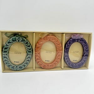 May include: A set of three oval picture frames in teal, peach, and purple, each featuring an ornate embossed border and a ribbon loop. The frames are displayed in a rectangular box, with each frame showing the text 2 1/2 in x 3 3/4 in.