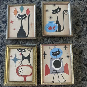 May include: Four rectangular gold-toned metal frames containing illustrations of black cats. The prints feature mid-century motifs including diamonds, a goldfish bowl, a red abstract shape, and a cat in a space helmet riding a rocket ship.