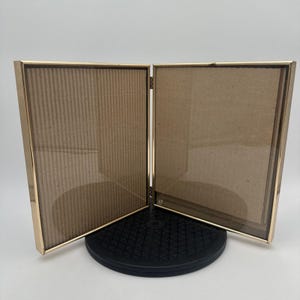 May include: A double-hinged picture frame featuring two rectangular panels with thin gold-toned metal borders. The frames are connected by a central hinge, allowing them to stand upright. Each panel contains a clear glass front and a brown backing.