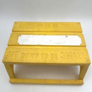 May include: A yellow plastic step stool featuring the embossed text STEP UP TO BE TALL on the top edge and SIT DOWN TO BE SMALL on the bottom edge. A rectangular silver-colored metal plate is centered horizontally between the two lines of text.