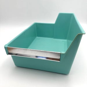 Puede incluir: Una bandeja de almacenamiento de plástico turquesa con un borde de metal plateado. La bandeja tiene forma rectangular con una parte trasera elevada y un fondo acanalado. La bandeja está vacía y sobre una superficie blanca.