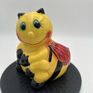 May include: A ceramic bee figurine with a glossy yellow body, black stripes, and black antennae. The bee has a smiling face with blue eyes and red cheeks. The wings are red with black detailing. The figurine is sitting on a black, textured surface.