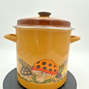May include: Mustard yellow enamel pot with a brown lid and handles. The pot features a whimsical mushroom design in orange, brown, and yellow. A vintage-style kitchenware item.