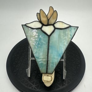 May include: A stained glass night light featuring a triangular shape with light blue and white marbled glass panels. The top section displays a stylized flower design in cream and white glass, all held together by dark metal solder lines.