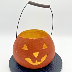 May include: A round orange jack-o'-lantern bucket with a textured surface, featuring cut-out triangular eyes, a triangular nose, and a jagged mouth. The bucket includes a thin black wire handle with a dark brown cylindrical grip at the top.