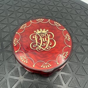 Può includere: Un compatto rotondo rosso con un bordo decorativo dorato e un monogramma dorato delle lettere "D" e "B" con una corona. Il compatto ha una finitura lucida e un bordo decorativo.