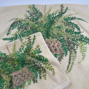 May include: Two cream-colored bath towels with a fern design. The towels feature a detailed print of a hanging fern basket with green and brown leaves. The towels have a soft, textured appearance.