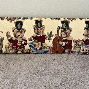 May include: A rectangular tapestry featuring five teddy bears in marching band uniforms. The bears are in red jackets and black hats, each holding a different instrument or object. The background is cream-colored with pinecone and greenery borders.