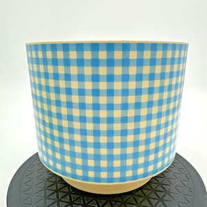 May include: A cylindrical planter featuring a light blue and cream-colored gingham check pattern. The vessel has a smooth, uniform exterior surface with a consistent grid design throughout, and it sits on a slightly recessed, solid cream-colored base.