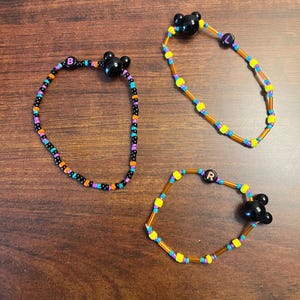 Könnte beinhalten: Drei Perlenarmbänder mit schwarzen Mickey-Maus-Kopf-Anhängern. Die Armbänder sind aus schwarzen, orangefarbenen, gelben und violetten Perlen gefertigt. Die Armbänder sind mit den Buchstaben "B", "L" und "R" gekennzeichnet.