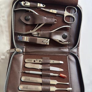 Pode incluir: Um kit de manicure em couro castanho com várias ferramentas de aço inoxidável, incluindo cortadores de unhas, tesouras e empurradores de cutículas. O kit está aberto, revelando as ferramentas cuidadosamente dispostas em um estojo com zíper. A marca "Bite" é visível.
