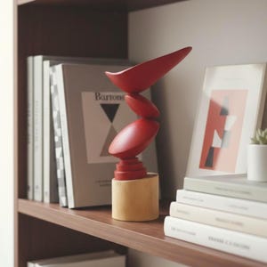 Peut inclure: Une sculpture abstraite moderne, rouge et bois naturel. Elle présente des formes arrondies empilées sur une base cylindrique. Exposée sur une étagère en bois avec des livres et des œuvres encadrées.