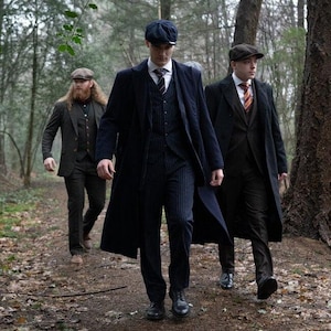Puede incluir: Tres personas con trajes de estilo vintage y gorras planas caminan por un sendero forestal. Los trajes son oscuros, con rayas y colores lisos. Cada persona lleva un abrigo largo, evocando una estética clásica e histórica.