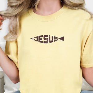 May include: A light yellow t-shirt with the word "JESUS" in a fish-shaped design. The shirt is a soft, comfortable material. The word "BUTTER" is in the upper right corner.