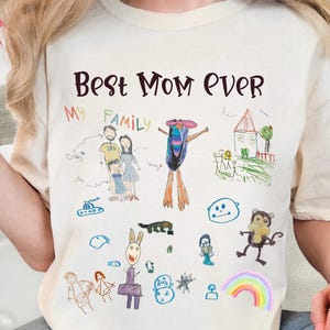 Può includere: T-shirt color avorio con la scritta "Best Mom Ever" e un colorato disegno per bambini. Il disegno include una famiglia, una casa, un arcobaleno e altri elementi fantasiosi. La maglietta ha un girocollo e maniche corte.