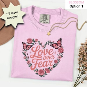 Op de afbeelding: Lichtroze T-shirt met een hartvormig bloemenontwerp en de tekst "Love over Fear". Het ontwerp bevat roze rozen, vlinders en tekst. Een gouden sterhangerketting ligt op het shirt.