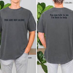 May include: Pepper-colored t-shirt with the text "YOU ARE NOT ALONE" on the front. The back of the shirt reads "You can talk to me, I'm here to help." The shirt is a short-sleeved, casual style.