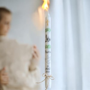 Puede incluir: Una vela de bautizo blanca encendida. La vela está decorada con una cruz dorada, hojas verdes, el nombre "Léo", la palabra "Bautismo", una paloma y la fecha "12 Abril 2026". Un lazo de cuerda está atado en la base.