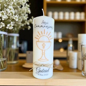 Pode incluir: Uma vela branca em forma de pilar com o texto "1ere Communion" e um desenho de cálice. A vela é decorada com um laço de barbante e o nome "Gabriel" e a data "15 Juin 2025".