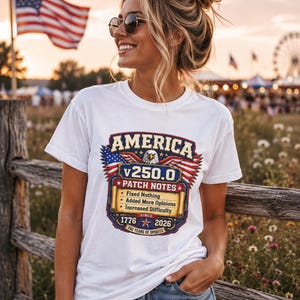 Op de afbeelding: Een wit t-shirt met een grafisch ontwerp met de tekst AMERICA v250.0 PATCH NOTES, met de vermelding Fixed Nothing, Added More Opinions en Increased Difficulty, met de data 1776 en 2026, een adelaar en een Amerikaans vlagmotief.