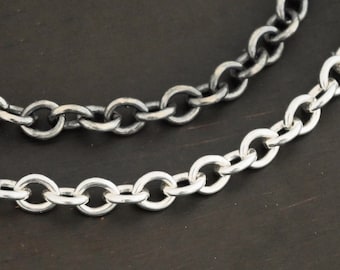 May include: Two silver chains, one is darker than the other, laid out on a dark brown wooden surface.