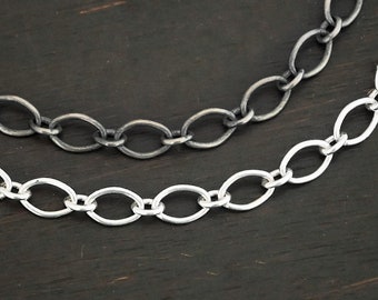 May include: Two silver chains, one oxidized and one polished, lay on a dark wood surface. The chains are both oval link chains.