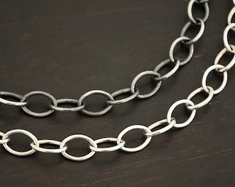May include: Two silver chains, one oxidized and one polished, lay on a dark wood surface. The chains are both oval link chains.