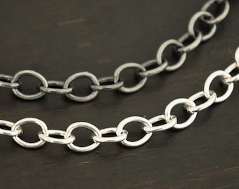 May include: Two silver chains, one oxidized and one polished, lay on a dark wood surface. The chains are both oval-linked and of different thicknesses.