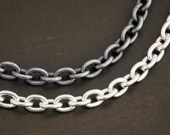 May include: Two silver chains, one black and one silver, are laid out on a dark brown wooden surface. The chains are both oval-linked.