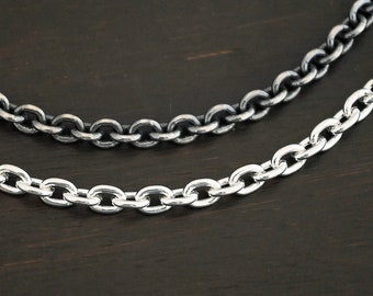 May include: Two silver chains, one oxidized and one polished, lay on a dark wood surface. The chains are both made of oval links.