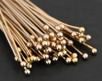 May include: A collection of gold-colored head pins with small, round ball ends. The pins are clustered together, with some showing a brighter, more polished finish. The background is a solid, dark color.