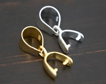 May include: Two pendant bails, one gold and one silver, are shown on a dark wood surface. The bails are designed to hold a pendant charm.