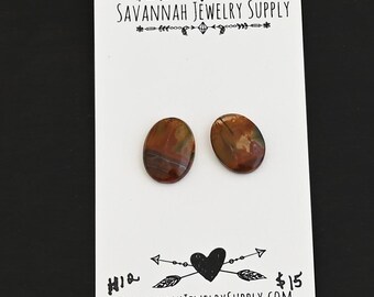 May include: Two oval-shaped, polished gemstone earrings in shades of brown, red, and green, displayed on a white card. The card features the text "SAVANNAH JEWELRY SUPPLY" and a decorative heart and arrow design.